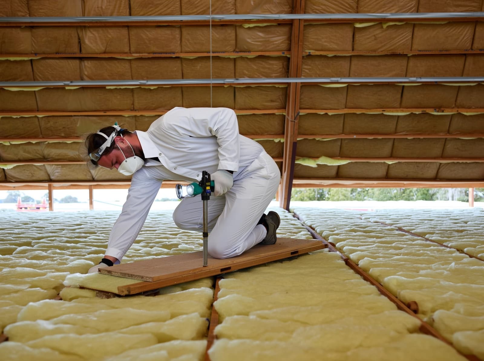 Installer in protective gear laying batts across the joists of a roof space